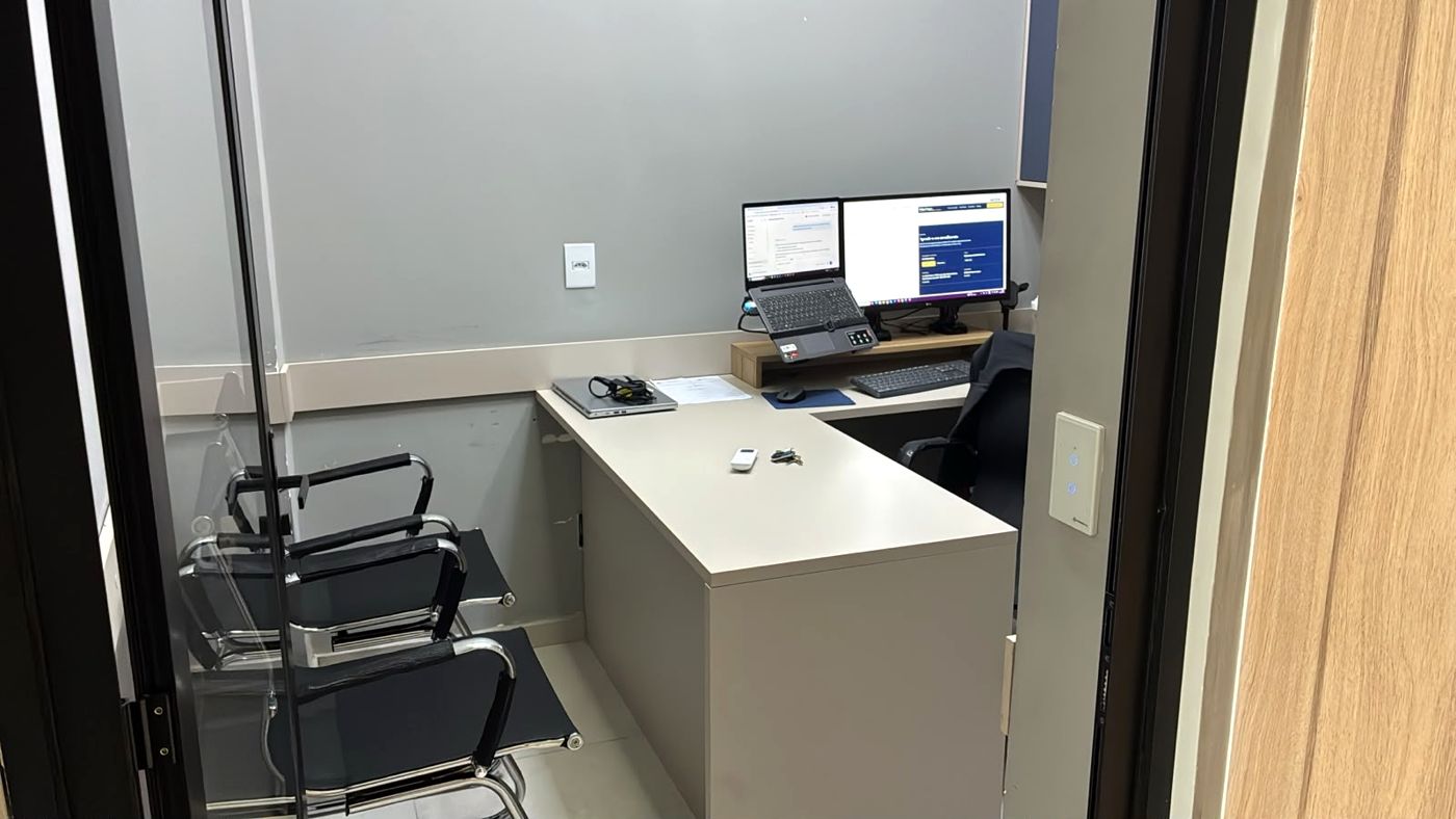 Sala de atendimento com mesa em L, cadeiras para clientes e dois monitores