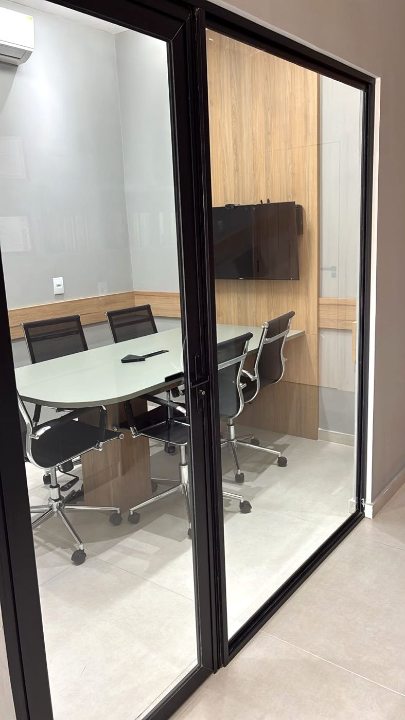 Sala de reunião com mesa oval, cadeiras e televisão, vista pela porta de vidro